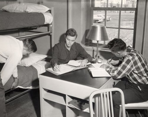 Three Vintage college students at desk in dorm room two are studying while third one is setting his bed. 
