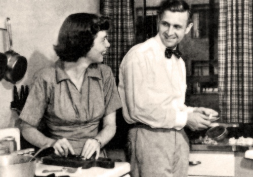 An old black and white photo of a man and woman preparing dinner in a kitchen.