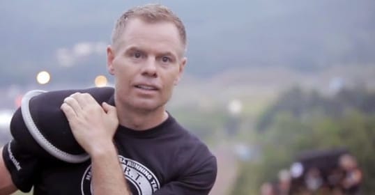 A man wearing a black shirt is holding a boxing bag while listening to the Spartan Up podcast.