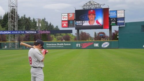 A manly baseball player holding a bat in front of a large screen.