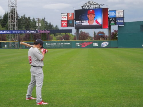 A manly baseball player holding a bat in front of a large screen.