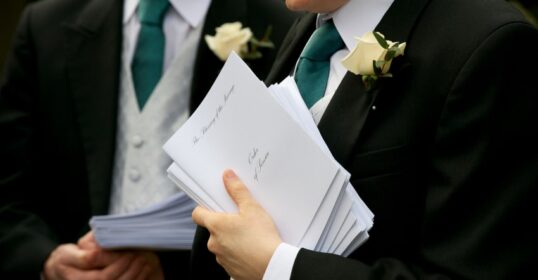 Two ushers in tuxedos holding papers.