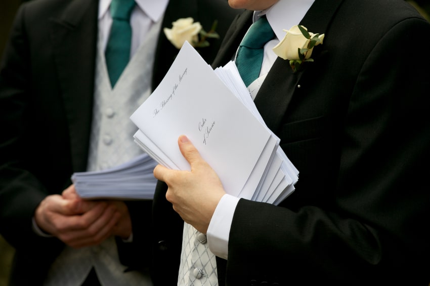 Two ushers in tuxedos holding papers.