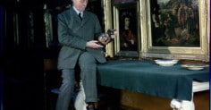 A man in a suit sitting at a table in front of paintings, appearing to invest.