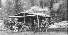 A group of manly men in front of a log cabin.