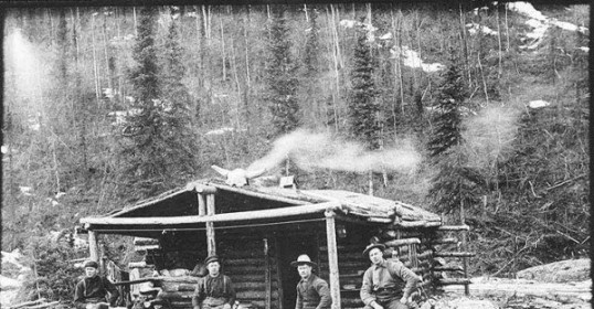 A group of manly men in front of a log cabin.