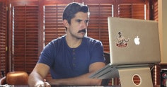 A man, working at a desk with a laptop and a mustache.