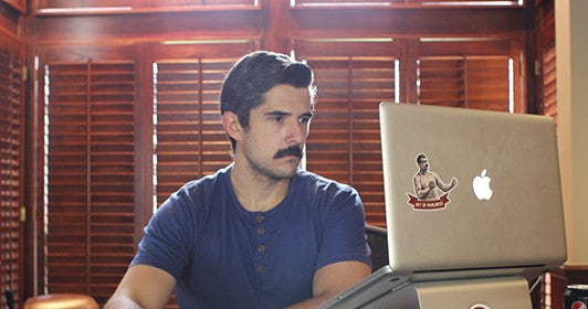 A man, working at a desk with a laptop and a mustache.