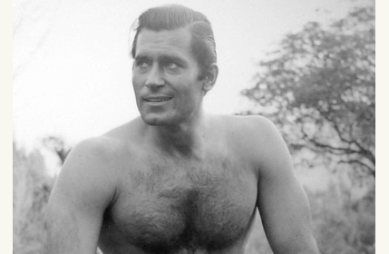 A shirtless man in a black and white photo exudes masculinity and showcases his muscular physique.