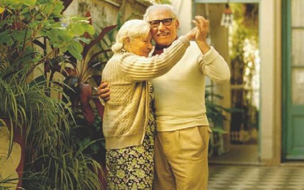 Old couple dancing at home with plants at the back.