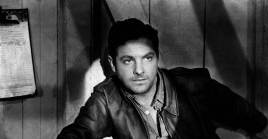 A man in a stylish leather jacket leaning against a wall.