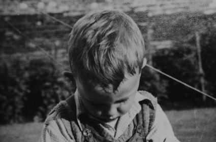 A kid in overalls having fun with a bowl of mud.