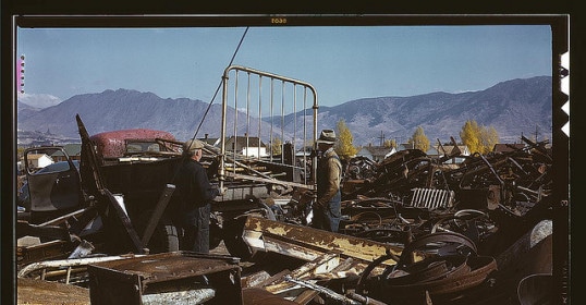 A group of men standing next to a pile of scrap metal, possibly looking to make money.