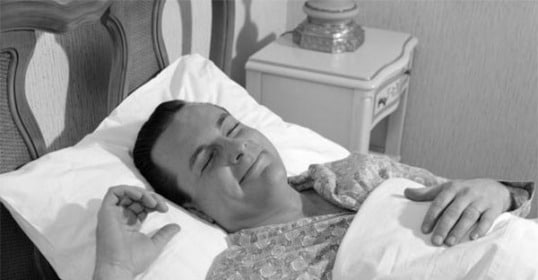 A man lying in bed with a cigarette, seemingly asleep.