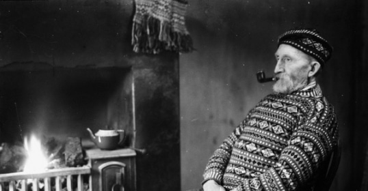 A man seated by the fireplace, puffing on a pipe.