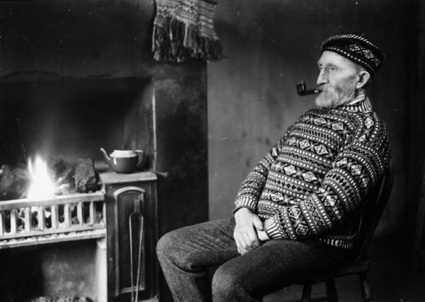 A man seated by the fireplace, puffing on a pipe.