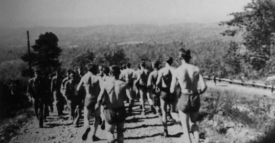 During WWII, a group of men is running on a dirt road as part of their workout regimen.