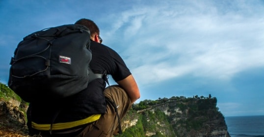 A man with a backpack sitting on a cliff overlooking the ocean, contemplating the vast world before him.