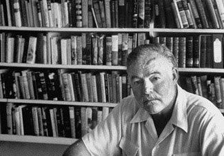 A black and white photo of a man sitting in front of a bookshelf, reminiscent of famous men like Ernest Hemingway.