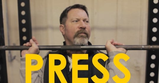 A man holding a barbell with the words press on it is demonstrating an overhead press in the video.
