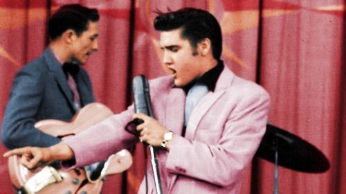 A man wearing a pink suit is singing into a microphone, resembling Elvis Presley.