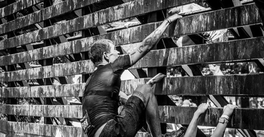 A man training for an obstacle race, dominating a wooden wall in a black and white photo.