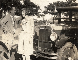 A man and a woman standing next to an old car as they potentially begin a pick-up scene.