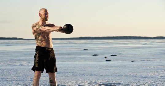 A man with tattoos showcasing his power swings a kettlebell in the snow.