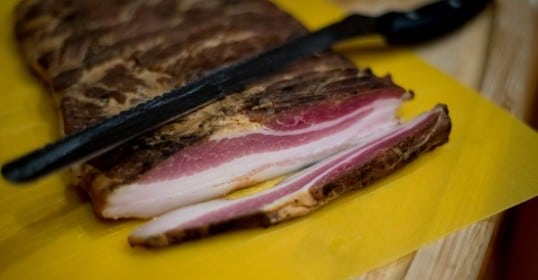 A piece of bacon on a cutting board, ready to be sliced with a knife.