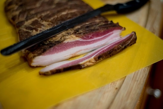 A piece of bacon on a cutting board, ready to be sliced with a knife.