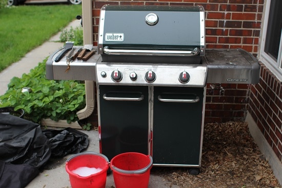 A clean green gas grill with a red bucket beside it.