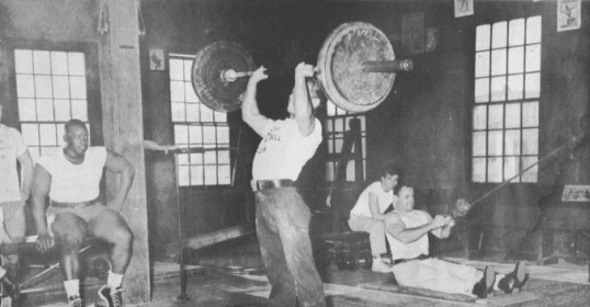 A black and white photo of a group of people in a gym, with some using barbells.