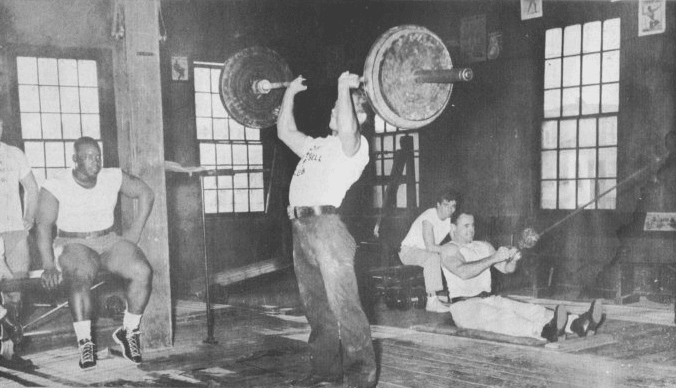 A black and white photo of a group of people in a gym, with some using barbells.