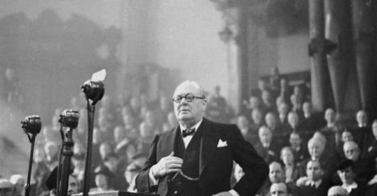 An old photo of a man in a suit standing in front of a crowd, reminiscent of Winston Churchill's public speaking.