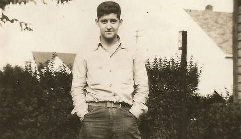A young man with his hands in his pockets stands in front of a house in this old photo.