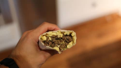 A person holding up a burrito with meat and eggs, perfect for saving time or make-ahead meals.