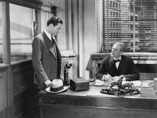 Two men standing at a desk discussing how to fire someone.