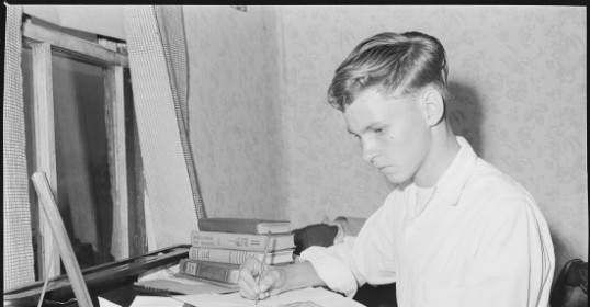 A young man writing at a desk for his college application essay.