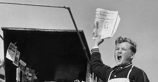 A boy keeping up with the news, holding a newspaper in front of a truck.