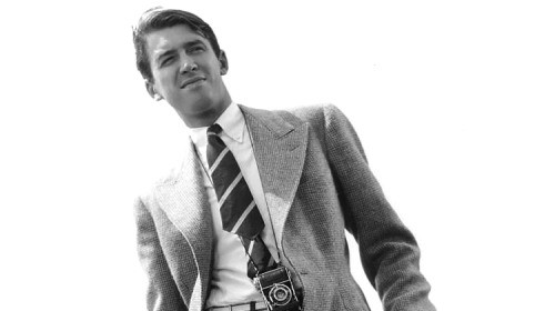 A stylish man in a suit leaning against a fence.
