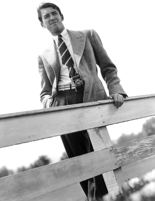 A stylish man in a suit leaning against a fence.