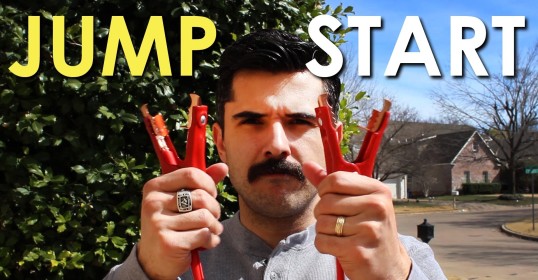 A man with a mustache is holding a pair of jumper cables next to a car he is about to jump start.