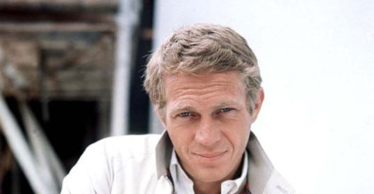 A man in a white jacket leaning against a wall, showcasing his style staples.