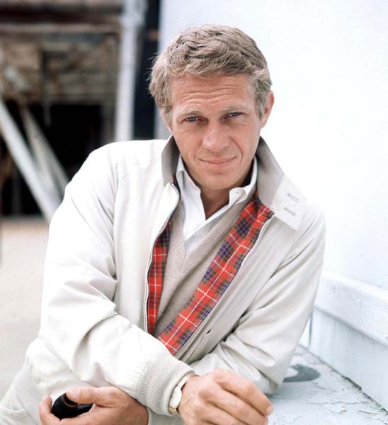 A man in a white jacket leaning against a wall, showcasing his style staples.