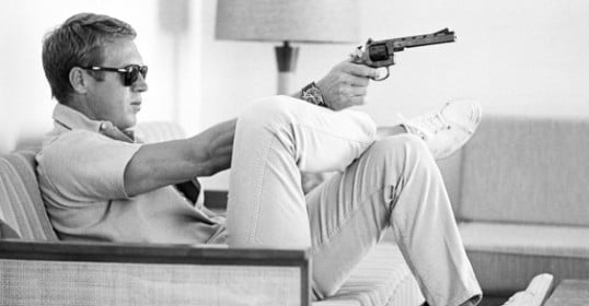 A man sitting on a couch holding a gun, channeling Steve McQueen for style inspiration in khakis.