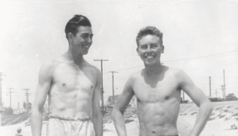 Two friends in swim trunks standing on the beach.