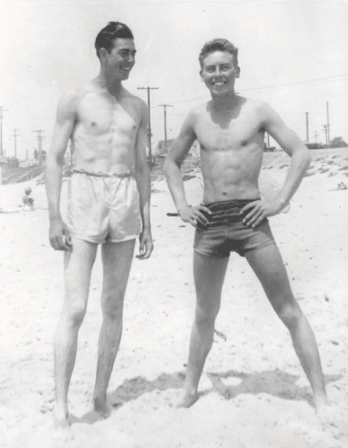 Two friends in swim trunks standing on the beach.