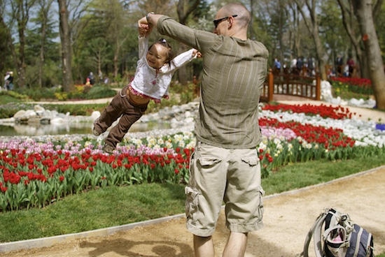 Dad swinging little girl by arms at botanical garden with bag placed on the floor.