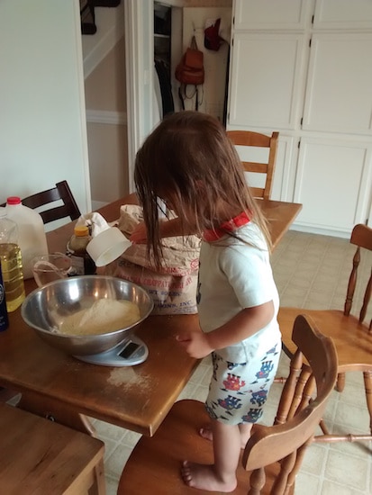 Little girl pouring flour into mixing bowl making bread while standing on a chair.