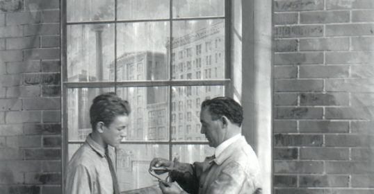 Two men standing in front of a table, one displaying subservience.